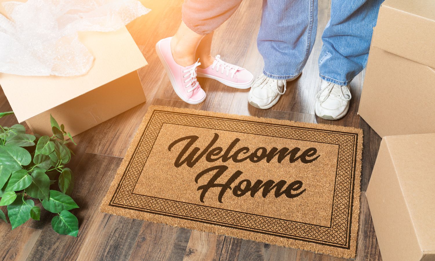 Close-up of two people standing near a "Welcome Home" closing gift doormat with unpacked boxes and a plant on a wooden floor.